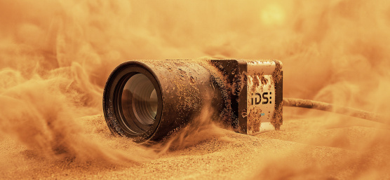Industrial camera partly buried in sand, surrounded by swirling sand.