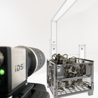 IDS uEye CP industrial camera pointing at a pallet cage with metal parts, while a modern lighting and scanning unit can be seen in the background