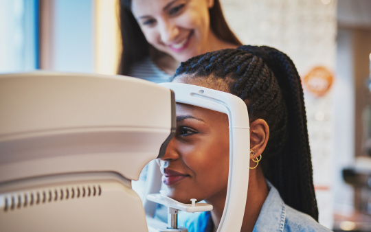 Woman looking through an ophthalmology device, optician standing behind her.