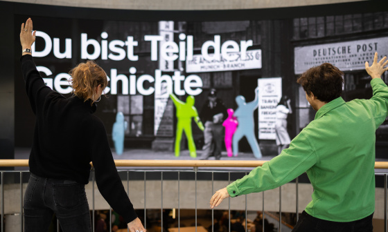 Two people are posing in front of a large interactive display wall featuring colorful silhouettes
