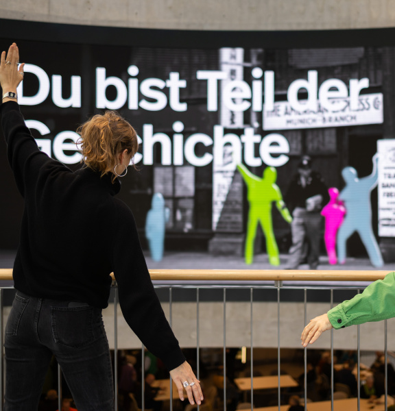 Two people are posing in front of a large interactive display wall featuring colorful silhouettes