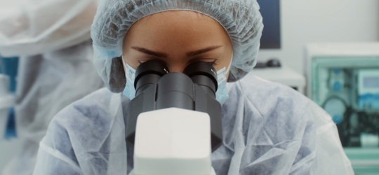 A woman in protective clothing looks into a microscope.