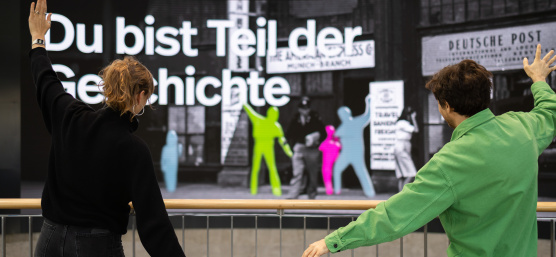 Two people are posing in front of a large interactive display wall featuring colorful silhouettes
